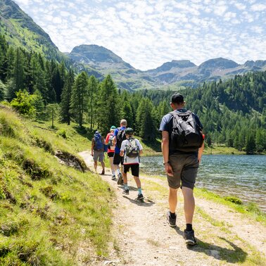 „Kinder und Erwachsene wandern entlang des Duizitzkarsees durch die alpine Landschaft mit Wald und Bergen.“ | © TVB/Laura Passenbrunner